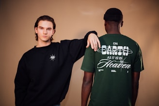 Two individuals are in a neutral-toned setting. One person, facing forward, wears a black long-sleeved shirt with a white emblem on the chest. The other individual, with their back to the camera, sports a green t-shirt with white text that reads 'ON EARTH AS IT IS IN HEAVEN'. Both people have a relaxed posture.