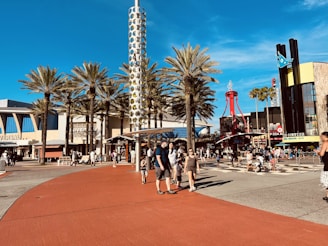 A bustling outdoor plaza featuring palm trees, a tall cylindrical structure with circular patterns, and crowds of people walking and gathering. The area includes signage for Universal Studios and various restaurants. The scene is lively with clear blue skies overhead.