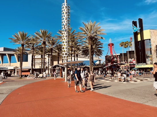 A bustling outdoor plaza featuring palm trees, a tall cylindrical structure with circular patterns, and crowds of people walking and gathering. The area includes signage for Universal Studios and various restaurants. The scene is lively with clear blue skies overhead.