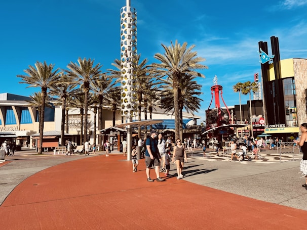A bustling outdoor plaza featuring palm trees, a tall cylindrical structure with circular patterns, and crowds of people walking and gathering. The area includes signage for Universal Studios and various restaurants. The scene is lively with clear blue skies overhead.