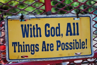 A rectangular yellow sign with a blue border displays the phrase 'With God, All Things Are Possible!' in bold, capitalized blue letters. The sign is mounted on a red metal mesh frame, and the background shows some greenery, indicating an outdoor setting.