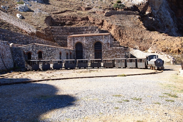 A historic stone building is situated at the base of a rocky hillside, featuring large arched doorways. In front of the building, there is an old industrial train with a series of carts, likely used for mining or transporting materials. The surrounding terrain is dry and rugged, with patches of grass and gravel.