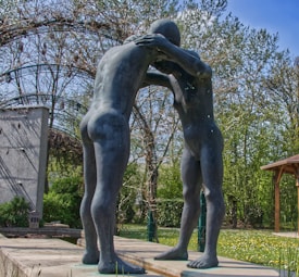 Two intertwined human-like sculptures are positioned face-to-face on a platform. The surrounding area includes greenery such as trees and bushes, as well as a structure with a wooden roof and an art piece arching behind the sculptures.