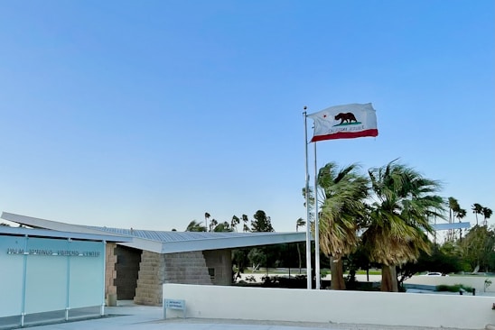 A clear blue sky frames a modern building with a flat roof labeled 'Palm Springs Visitors Center.' A California state flag waves atop a pole in the foreground. Tall palm trees sway gently beside the building, suggesting a light breeze.