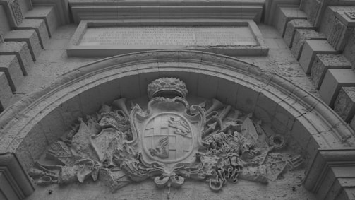 A detailed stone carving featuring an ornate coat of arms, set above an arched stone structure. The crest includes a shield with heraldic symbols and is surrounded by intricate foliage and decorative flourishes. Above the carving, an inscription is engraved into the stone.