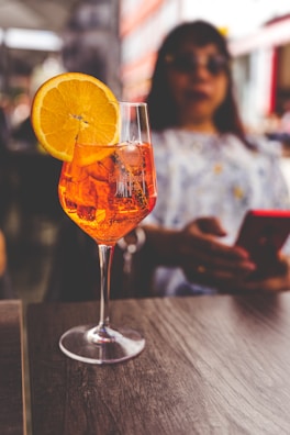 A glass of Aperol Spritz with a slice of orange sits on a wooden table. In the background, a person is holding a smartphone. The setting appears to be an outdoor cafe, with blurred buildings in the distance.