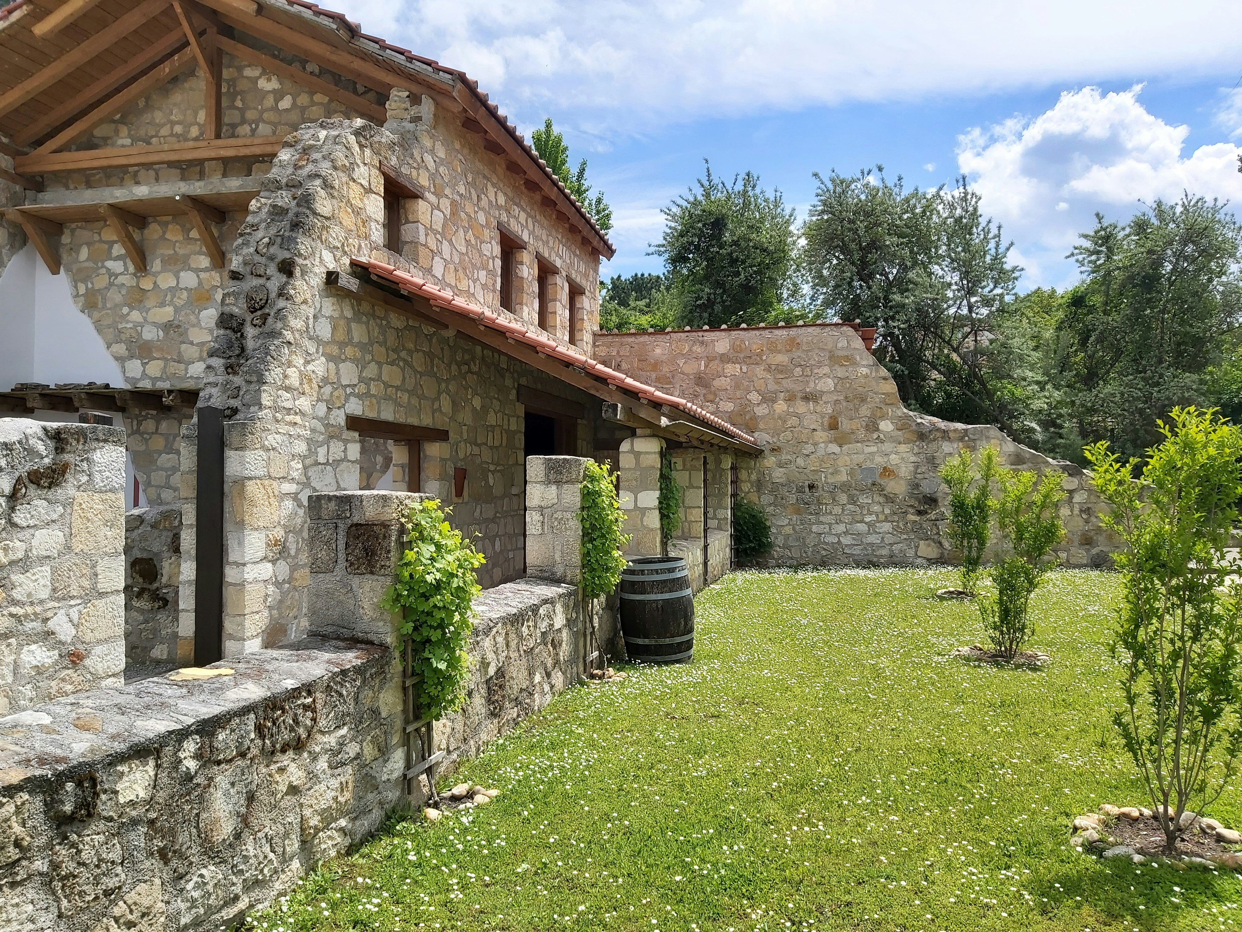 A rustic stone house with a tiled roof is set amidst a green garden. The architecture features wooden beams and weathered stone walls, surrounded by lush trees and grass. A wooden barrel and small shrubs accentuate the peaceful, natural environment.