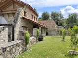 A rustic stone house with a tiled roof is set amidst a green garden. The architecture features wooden beams and weathered stone walls, surrounded by lush trees and grass. A wooden barrel and small shrubs accentuate the peaceful, natural environment.