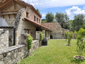 A rustic stone house with a tiled roof is set amidst a green garden. The architecture features wooden beams and weathered stone walls, surrounded by lush trees and grass. A wooden barrel and small shrubs accentuate the peaceful, natural environment.