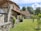 A rustic stone house with a tiled roof is set amidst a green garden. The architecture features wooden beams and weathered stone walls, surrounded by lush trees and grass. A wooden barrel and small shrubs accentuate the peaceful, natural environment.