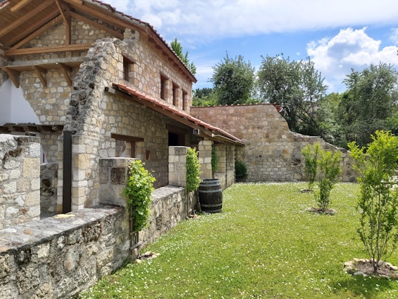 A rustic stone house with a tiled roof is set amidst a green garden. The architecture features wooden beams and weathered stone walls, surrounded by lush trees and grass. A wooden barrel and small shrubs accentuate the peaceful, natural environment.