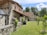 A rustic stone house with a tiled roof is set amidst a green garden. The architecture features wooden beams and weathered stone walls, surrounded by lush trees and grass. A wooden barrel and small shrubs accentuate the peaceful, natural environment.