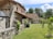 A rustic stone house with a tiled roof is set amidst a green garden. The architecture features wooden beams and weathered stone walls, surrounded by lush trees and grass. A wooden barrel and small shrubs accentuate the peaceful, natural environment.