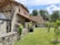 A rustic stone house with a tiled roof is set amidst a green garden. The architecture features wooden beams and weathered stone walls, surrounded by lush trees and grass. A wooden barrel and small shrubs accentuate the peaceful, natural environment.