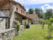 A rustic stone house with a tiled roof is set amidst a green garden. The architecture features wooden beams and weathered stone walls, surrounded by lush trees and grass. A wooden barrel and small shrubs accentuate the peaceful, natural environment.