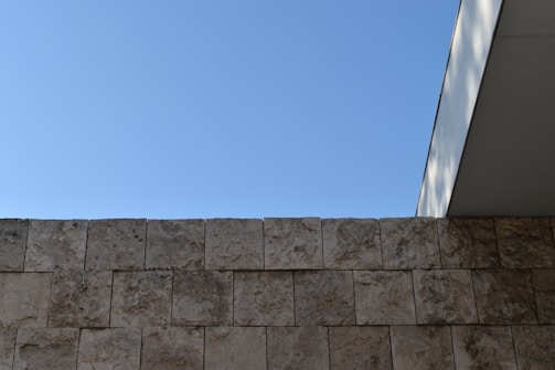 A minimalist architectural composition featuring a stone wall made of rectangular blocks in the foreground. Above, a clear blue sky is visible with the edge of a modern building's overhang entering the frame from the right, casting soft shadows.