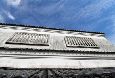 A traditional building facade with white plaster walls and wooden slatted windows. The roof features traditional curved tiles, and the sky is a clear blue with some clouds.