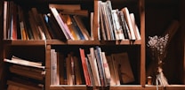 A wooden bookshelf filled with a variety of books, some leaning against each other while others are stacked. The shelves are lit warmly, creating a cozy atmosphere. To the right, a bundle of dried flowers is placed next to the books, adding a rustic touch.