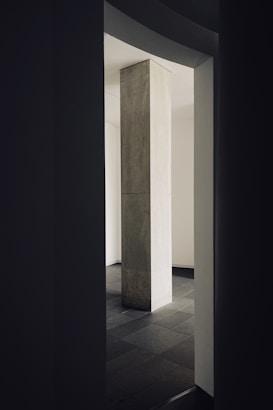 A tall rectangular pillar stands in the center of the image, surrounded by dark walls and a tiled floor. Light gently illuminates the pillar against a backdrop of muted tones.