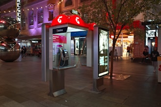 An outdoor public payphone booth is lit up with a distinctive red and white color scheme. The area features a large, stainless steel sculpture made of reflective spherical shapes nearby. People are walking and sitting around the open plaza area, which is brightly illuminated with neon signage and shop fronts. Trees and modern architecture add to the urban setting.