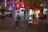 An outdoor public payphone booth is lit up with a distinctive red and white color scheme. The area features a large, stainless steel sculpture made of reflective spherical shapes nearby. People are walking and sitting around the open plaza area, which is brightly illuminated with neon signage and shop fronts. Trees and modern architecture add to the urban setting.