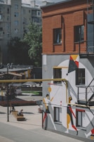 A modern urban setting with a red brick building featuring a bold, abstract mural painted on the side. The mural includes geometric shapes in black, white, red, yellow, and orange, creating a dynamic and vibrant visual. In the foreground, a wooden bench with people sitting adds a touch of everyday life to the scene. The backdrop includes additional buildings and greenery.
