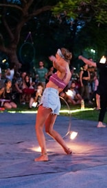 A woman is performing a fire dance in a park during the evening. She is wearing a purple crop top, light denim shorts, and a headband. In the background, there is a crowd of people watching the performance. Some are sitting while others are standing. It appears to be a relaxed gathering under trees with dim, ambient lighting.