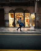 A person walks past a brightly lit clothing store with elegant dresses displayed in the windows. The storefront features an ornate design above the windows, and the sidewalk is lined with yellow traffic lines. The scene has a contrast of the warm interior light against the dim exterior street.