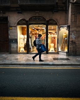 A person walks past a brightly lit clothing store with elegant dresses displayed in the windows. The storefront features an ornate design above the windows, and the sidewalk is lined with yellow traffic lines. The scene has a contrast of the warm interior light against the dim exterior street.