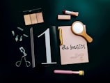 A flat lay arrangement featuring makeup items, including a highlighter palette, lipstick, eyelash curler, and a makeup brush. A small wooden hand mirror, quartz crystals, and a notebook with 'the basics' written on it are also present. These items are set against a dark background, conveying a sense of elegance and simplicity.