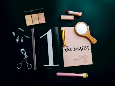 A flat lay arrangement featuring makeup items, including a highlighter palette, lipstick, eyelash curler, and a makeup brush. A small wooden hand mirror, quartz crystals, and a notebook with 'the basics' written on it are also present. These items are set against a dark background, conveying a sense of elegance and simplicity.