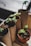 Several small seedlings with green leaves are planted in cardboard tubes and a single terracotta pot. The tubes resemble recycled toilet paper rolls, indicating an eco-friendly approach to gardening. The seedlings are placed in a wooden tray, and the focus is on their fresh green leaves emerging from the soil.