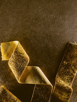 A curled, gold glitter ribbon is placed against a textured, dark background. The ribbon has a shiny and reflective surface, creating a contrast with the matte finish of the surrounding surface.