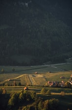 Sunlit green fields and wide roads within a peaceful farmhouse community surrounded by trees.