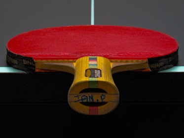 A table tennis paddle with a red rubber surface is placed upright on a dark table, displaying its wooden handle with colorful stripes.