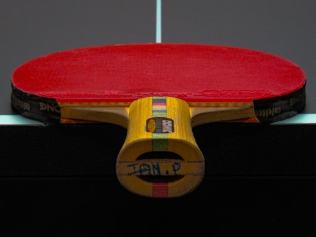 A table tennis paddle with a red rubber surface is placed upright on a dark table, displaying its wooden handle with colorful stripes.