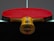 A table tennis paddle with a red rubber surface is placed upright on a dark table, displaying its wooden handle with colorful stripes.