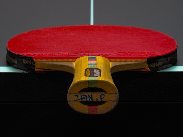 A table tennis paddle with a red rubber surface is placed upright on a dark table, displaying its wooden handle with colorful stripes.