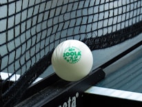 A close-up of a table tennis ball resting against the net on a table tennis table. The ball is branded with the words 'JOOLA 40+ FLIP' in green lettering. The net is tightly woven, and part of the table surface is visible.