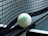 A close-up of a table tennis ball resting against the net on a table tennis table. The ball is branded with the words 'JOOLA 40+ FLIP' in green lettering. The net is tightly woven, and part of the table surface is visible.