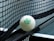 A close-up of a table tennis ball resting against the net on a table tennis table. The ball is branded with the words 'JOOLA 40+ FLIP' in green lettering. The net is tightly woven, and part of the table surface is visible.