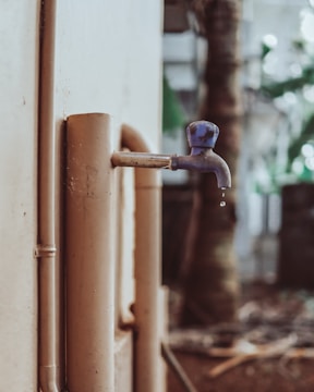 A close-up of an outdoor water faucet with a single droplet hanging from its spout. The faucet is attached to a beige-colored pipe, with a blurred natural background that includes trees and foliage.