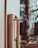 A close-up of an outdoor water faucet with a single droplet hanging from its spout. The faucet is attached to a beige-colored pipe, with a blurred natural background that includes trees and foliage.