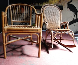 Two wooden chairs are placed on a red carpeted floor. One chair has a simple design with a slatted backrest, while the other is a decorative rocking chair with an intricate woven pattern on the backrest. In the background, there is a wall with abstract artwork depicting bird silhouettes.