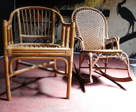 Two wooden chairs are placed on a red carpeted floor. One chair has a simple design with a slatted backrest, while the other is a decorative rocking chair with an intricate woven pattern on the backrest. In the background, there is a wall with abstract artwork depicting bird silhouettes.