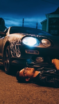 A person wearing glasses and a leather jacket is lying on a paved street at night, partially under a car with customized patterns. One of the car's headlights is on, casting a bright light.