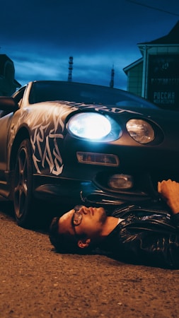 A person wearing glasses and a leather jacket is lying on a paved street at night, partially under a car with customized patterns. One of the car's headlights is on, casting a bright light.