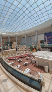 A spacious indoor area with a high, domed glass ceiling allowing natural light to illuminate the space. Below, there's a café or restaurant seating area with several tables and chairs arranged around a central food counter. The decor features large, intricate sculptures and art installations hanging from the ceiling. The floor is made of polished wood, and there's a sleek, curved edge with water features at the front.