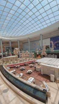 A spacious indoor area with a high, domed glass ceiling allowing natural light to illuminate the space. Below, there's a café or restaurant seating area with several tables and chairs arranged around a central food counter. The decor features large, intricate sculptures and art installations hanging from the ceiling. The floor is made of polished wood, and there's a sleek, curved edge with water features at the front.
