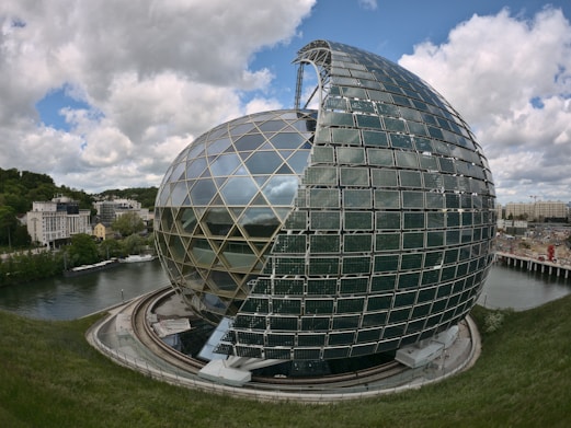 A large, modern spherical building with a partial open structure covered with solar panels sits beside a river. The building's glass and metal design reflects light, and surrounding it are green trees and urban structures. The sky is filled with scattered clouds, adding to the futuristic feel of the setting.